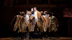Reuben Joseph playing the role of Alexander Hamilton is standing at the centre of the stage and is surrounded by other cast members of Hamilton the Musical, all dressed in brown and beige uniforms.