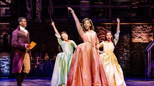 Trois femmes, les Schuyler Sisters, se tiennent ensemble sur scène, les bras en l'air, habillées dans de magnifiques robes d'époque de couleur pastel.