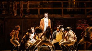 The actor who is playing Hamilton is standing in a long brown leather coat and is surrounded by members of the cast who are dancing around him.