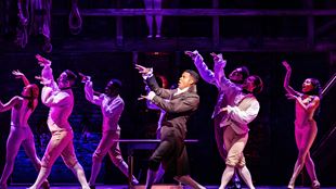 Les acteurs de la comédie musicale Hamilton dansent sur scène en costumes d'époque tandis qu'une lumière violette est tamisée sur la scène.