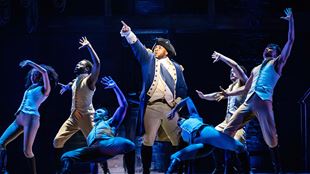 Thomas Vernal playing the role of Georges Washington is standing at the centre of the stage and is surrounded by other cast members of Hamilton the Musical, all dressed in blue and beige uniforms.