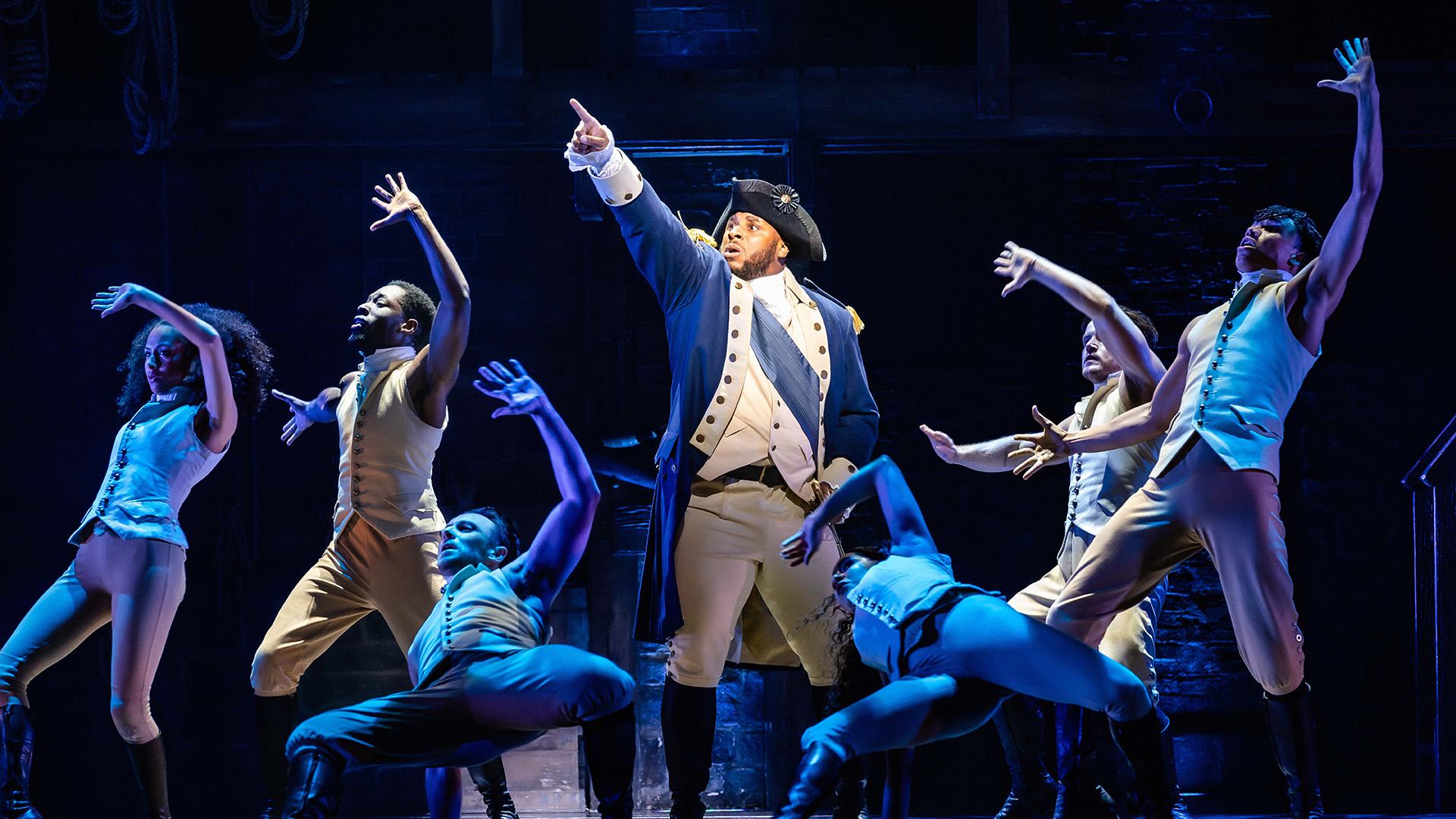 Allez voir la comédie musicale primée Hamilton, actuellement au Victoria Palace Theatre, et découvrez l'histoire du père fondateur des États-Unis. Image reproduite avec l'aimable autorisation de Raw PR/crédit photo Danny Kaan. Thomas Vernal, qui joue le rôle de Georges Washington, se tient au centre de la scène et est entouré d'autres membres de la troupe de la comédie musicale Hamilton, tous vêtus d'uniformes bleus et beiges.