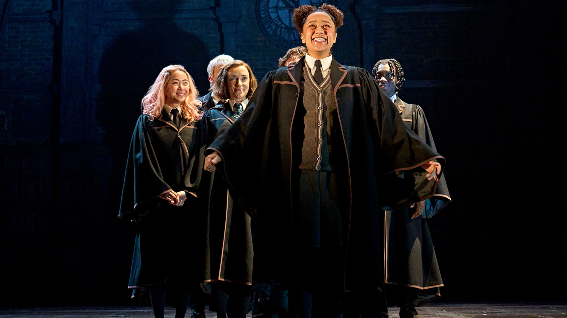 Rejoignez Harry et ses amis face à un nouveau défi dans Harry Potter et l'enfant maudit, une pièce magique actuellement au Palace Theatre. © London Theatre Direct L'actrice, qui incarne Rose Weasley dans Harry Potter et l'enfant maudit, porte l'uniforme de Poudlard et se tient fièrement sur scène, souriante, tandis que ses camarades de classe se tiennent derrière elle.