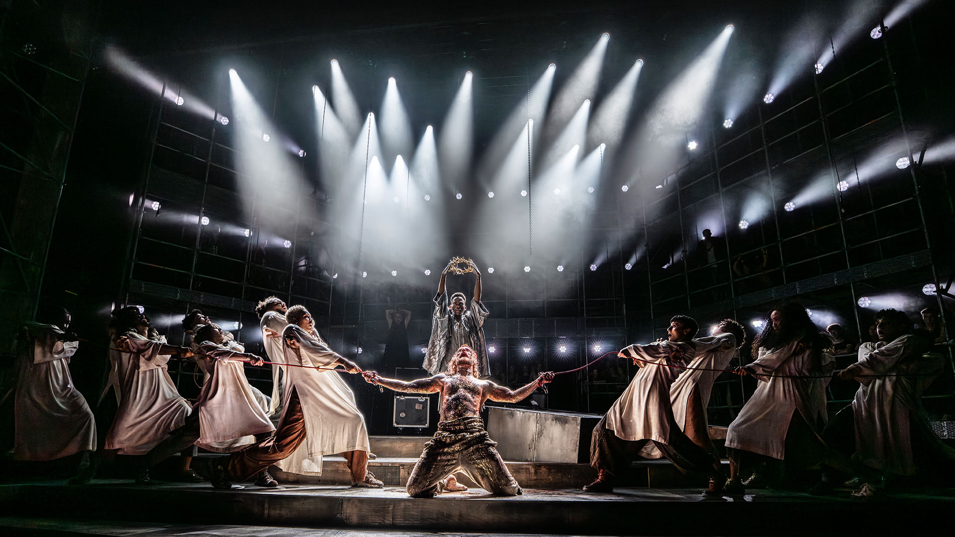 Watch the revival of Andrew Lloyd Webber's opera rock Jesus Christ Superstar, starring Sam Ryder. Photo credit: Johan Persson. The cast of Jesus Christ Superstar stand on stage, with Sam Ryder kneeling at the centre and raising his arms towards the air, surrounded by the other cast members.