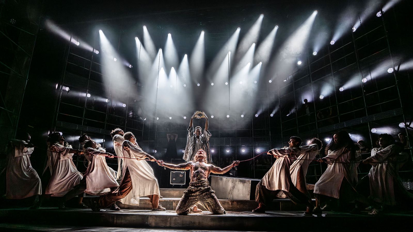 The cast of Jesus Christ Superstar stand on stage, with Sam Ryder kneeling at the centre and raising his arms towards the air, surrounded by the other cast members.