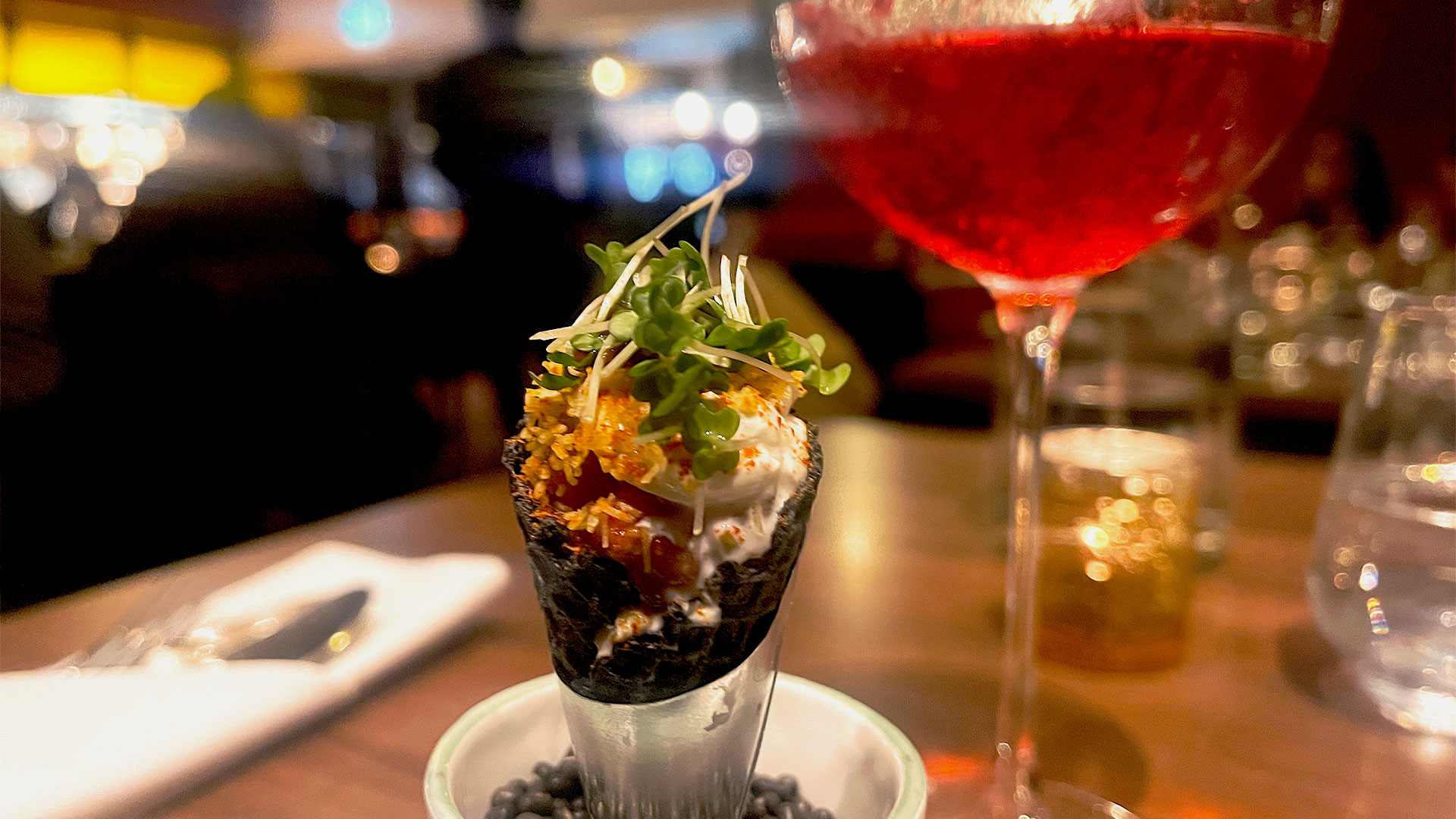 A red cocktail is served at the table alongside an amuse-bouche: a guava cone filled with tamarind chutney and raspberry sauce. This is part of Colonel Saab Trafalgar Square's Kinky Boots-themed menu.