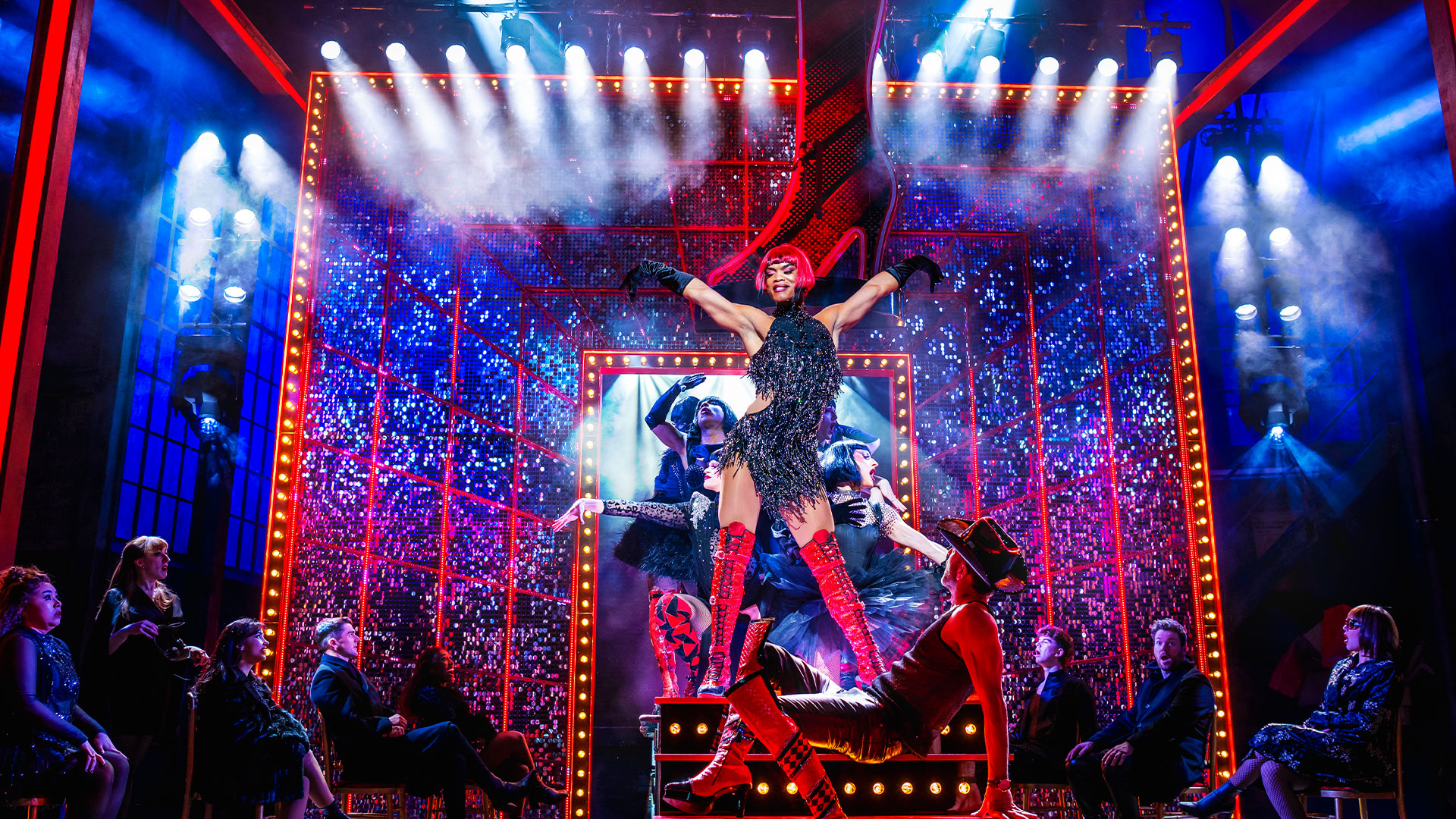 Johannes Radebe stands on stage wearing the infamous red-heeled boots, a red wig, and a sparkly black dress. A giant red heel is suspended above the stage.