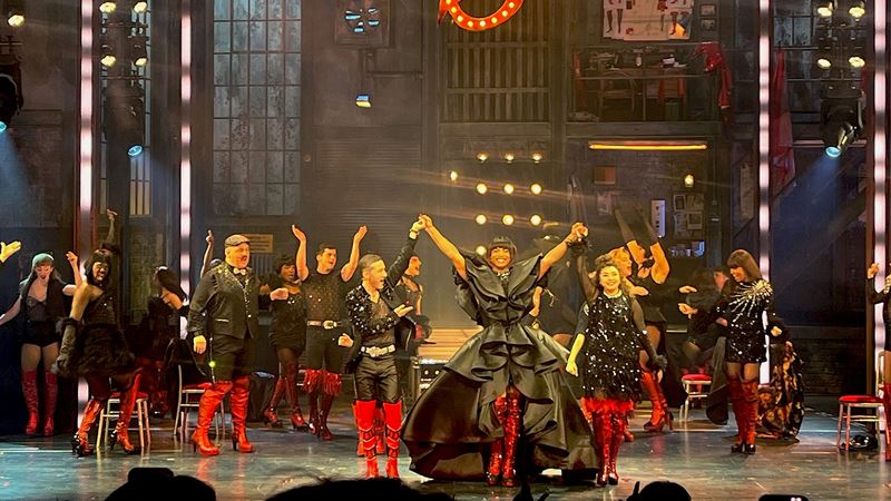 Johannes Radebe, wearing a flowing black gown and red-heeled boots, stands on stage with the cast of Kinky Boots at curtain call.