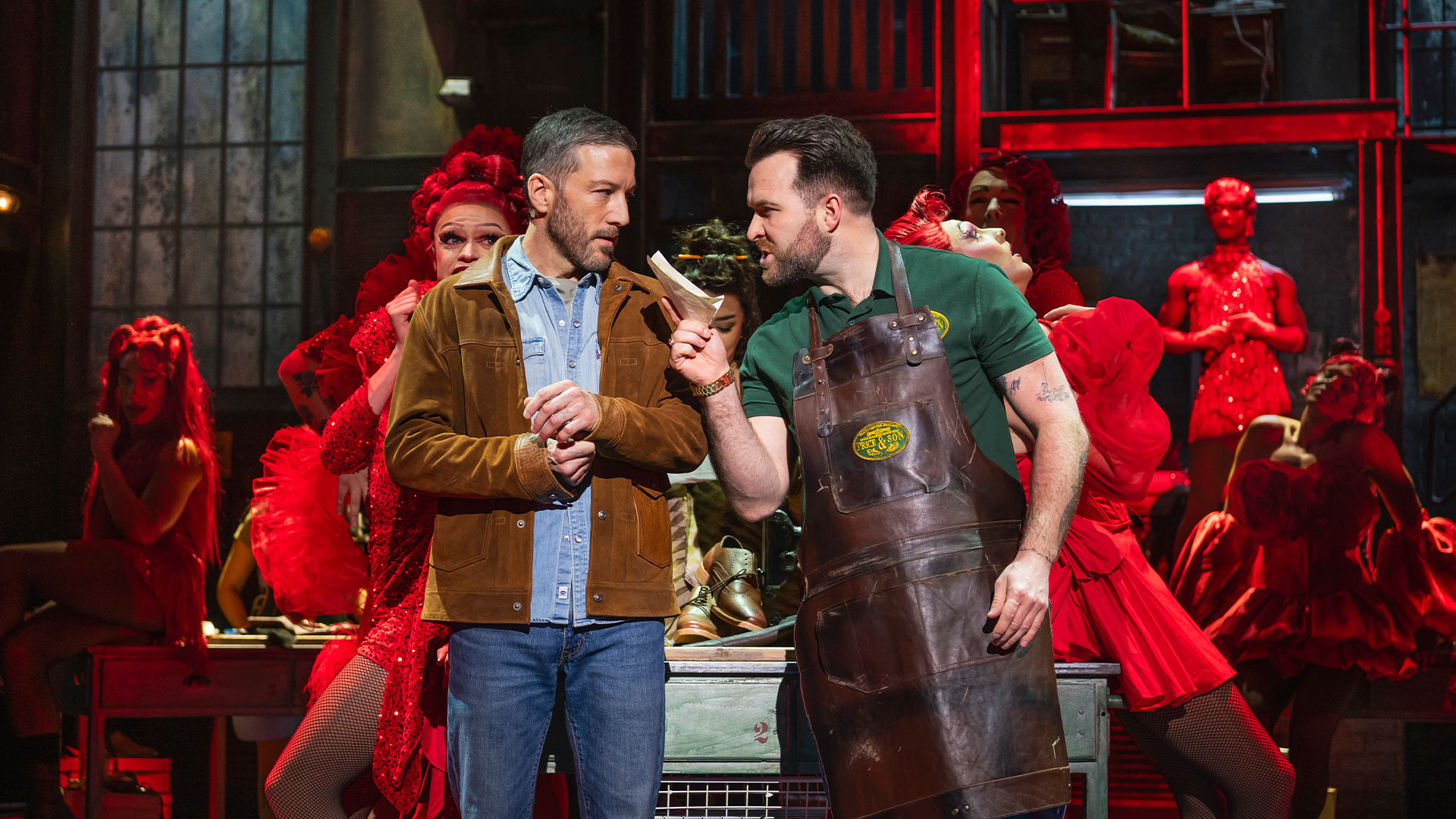 Watch the musical Kinky Boots and immerse yourself in a story that blends the struggles of factory workers with the fabulous world of drag queen performances. Photo credit: Matt Crockett. Matt Cardle and Billy Robert stand on the stage of the London Coliseum, with the Kinky Boots drag queen performers in red shadows behind them.
