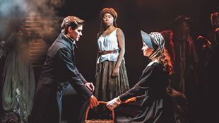 A man and a woman in period costume, looking at each other while holding a basket while a woman standing behind looks at them on a dark evening.