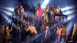 Les acteurs du spectacle Les Misérables sont debout sur la scène et lèvent les bras vers leurs compagnons d'armes.