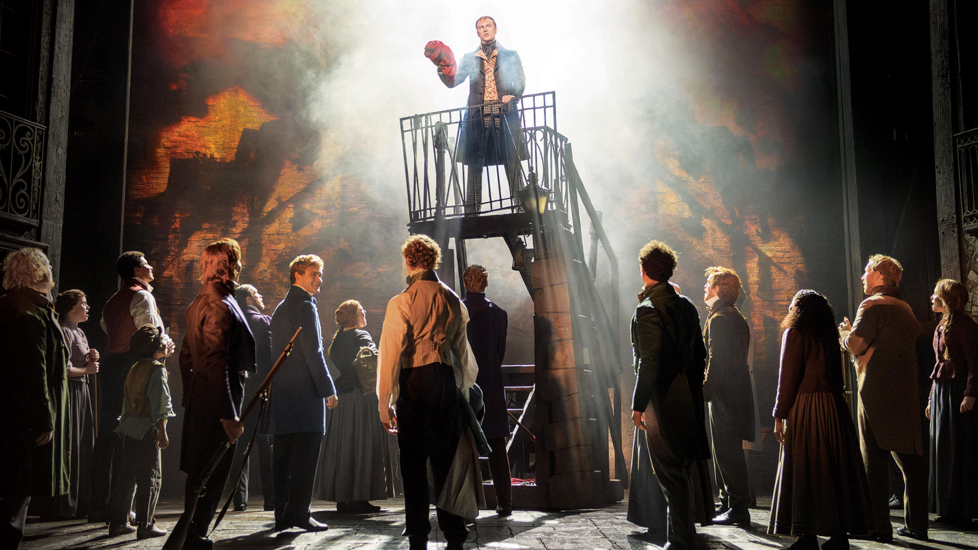 A man stands on a platform above a crowd of actors part of Les Misérables cast and makes a speech.