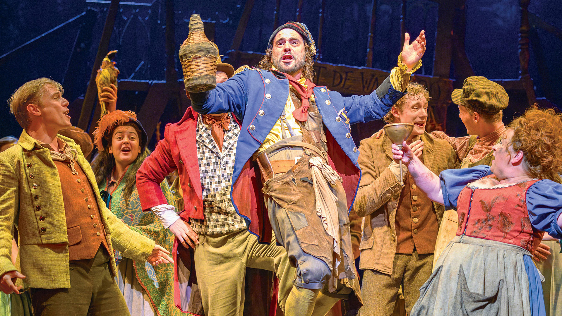 Découvrez l'adaptation théâtrale du roman à succès de Victor Hugo, Les Misérables, actuellement à l'affiche au Sondheim Theatre. Avec l'aimable autorisation de Story House PR/ Crédit photo : Deen Van Meer. Un homme portant un pantalon long bleu et marron de style militaire tient une bouteille et est entouré d'autres membres de la troupe des Misérables.