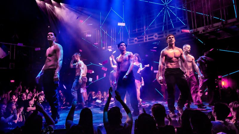 See Magic Mike Live on stage and get ready for a wild ride! Credit: Danny Kaan. Image courtesy of London Theatre Direct. The cast of Magic Mike Live is on stage in black trousers and shirtless with women in the audience with their hands up in the air and blue and red lights above them.