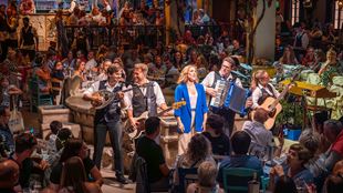 A woman in a blue jacket sings, accompanied by a band, surrounded by an audience seated at tables around the room at Mamma Mia! The Party.