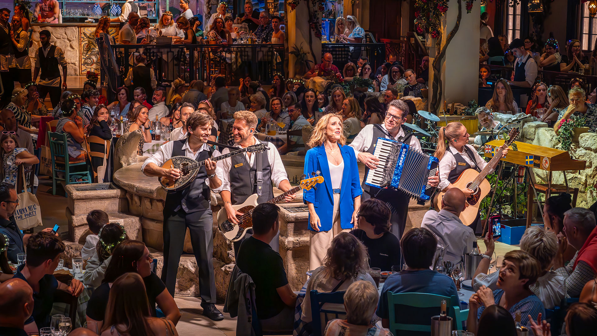 Let yourself be transported to sunny Greece for an evening with the immersive experience of Mamma Mia! The Party. Image courtesy of About Grace PR. Photo credit: Marc Brenner. Une femme en veste bleue chante, accompagnée d'un groupe de musiciens, entourée d'un public assis à des tables autour de la salle de Mamma Mia ! The Party.