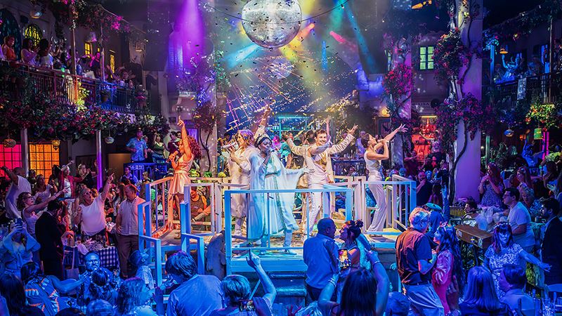 Mamma Mia! The Party cast members, dressed in sparkly white outfits, sing and dance on a small elevated stage as members of the audience join in.