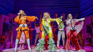 Drei Schauspielerinnen in bunten Kostümen aus der Disco-Ära tanzen auf der Bühne von Mamma Mia! Das Musical.