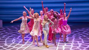 Women from the cast of Mamma Mia! are dressed in all shades of pink and dancing on the stage of the Novello Theatre.
