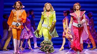 Three actresses dressed with colourful costumes from the disco era are dancing on the stage of MAMMA MIA! The Musical. 