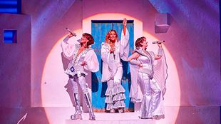 Three cast members of Mamma Mia dressed in silver outfits performing a song on stage under the spotlight