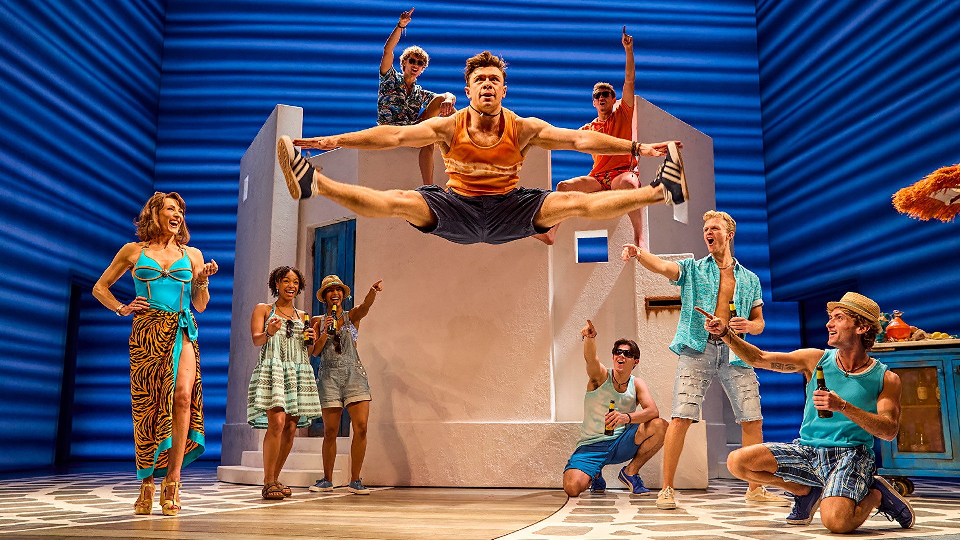 Kate Graham impresses in Tanya in MAMMA MIA! Image courtesy of Amanda Malpass/Brinkhoff & Mögenburg. The cast of Mamma Mia! The Musical is on stage in beach clothes, watching one of the dancers soar through high jumps with incredible energy.
