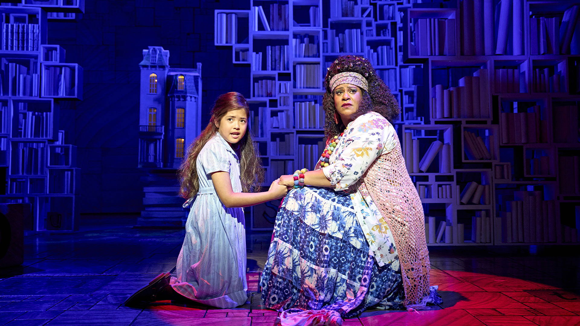 Matilda and the librarian stands on the stage of the Cambridge Theatre holding hands and looking at the audience with a worried look.