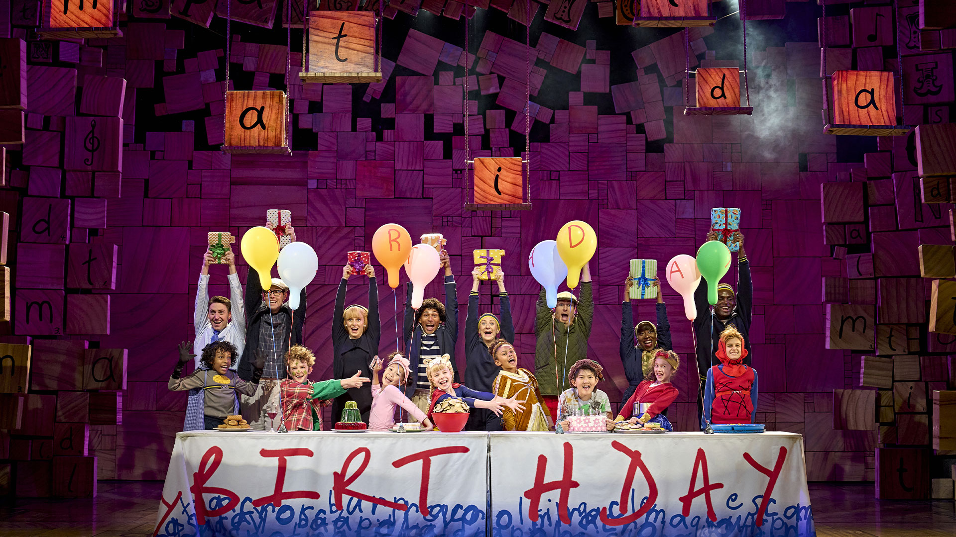 In Matilda the Musical, children and adults playing pupils and their parents stand behind a table covered with balloons and a tablecloth spelling out 'BIRTHDAY' in bright, bold red letters.