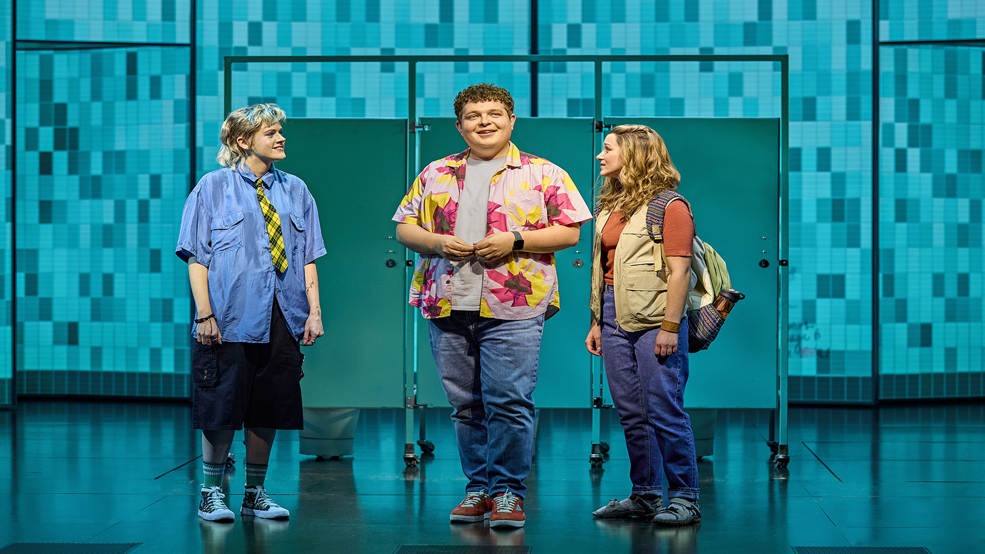 Elena Skye (Janis), Tom Xander (Damian) and Charlie Burn (Cady) in Mean Girls. Image courtesy of London Theatre Direct. Credits: Brinkhoff-Moegenburg Elena Skye, Tom Xander and Charlie Burn from the Mean Girls cast stand next to each other in front of three doors with a blue bathroom backdrop.