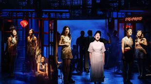 The production of Miss Saigon features a cast of performers standing on stage, with some of the female cast members wearing nightwear.