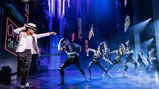 Myles Frost dances on stage, playing the iconic Michael Jackson in MJ the Musical, wearing the famous white hat and pointing to dancers all dressed in black bodysuits. 