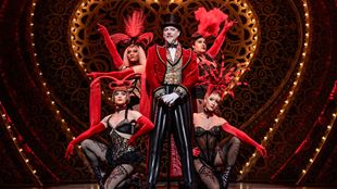 Harold Zidler, le propriétaire du Moulin Rouge, se tient sur le devant de la scène du Picadilly Theatre avec, derrière lui, quatre danseuses de cabaret vêtues de corsets en dentelle.