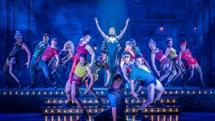 The cast of Moulin Rouge The Musical are on stage, wearing colourful outfits and dancing around the actress playing Satine.