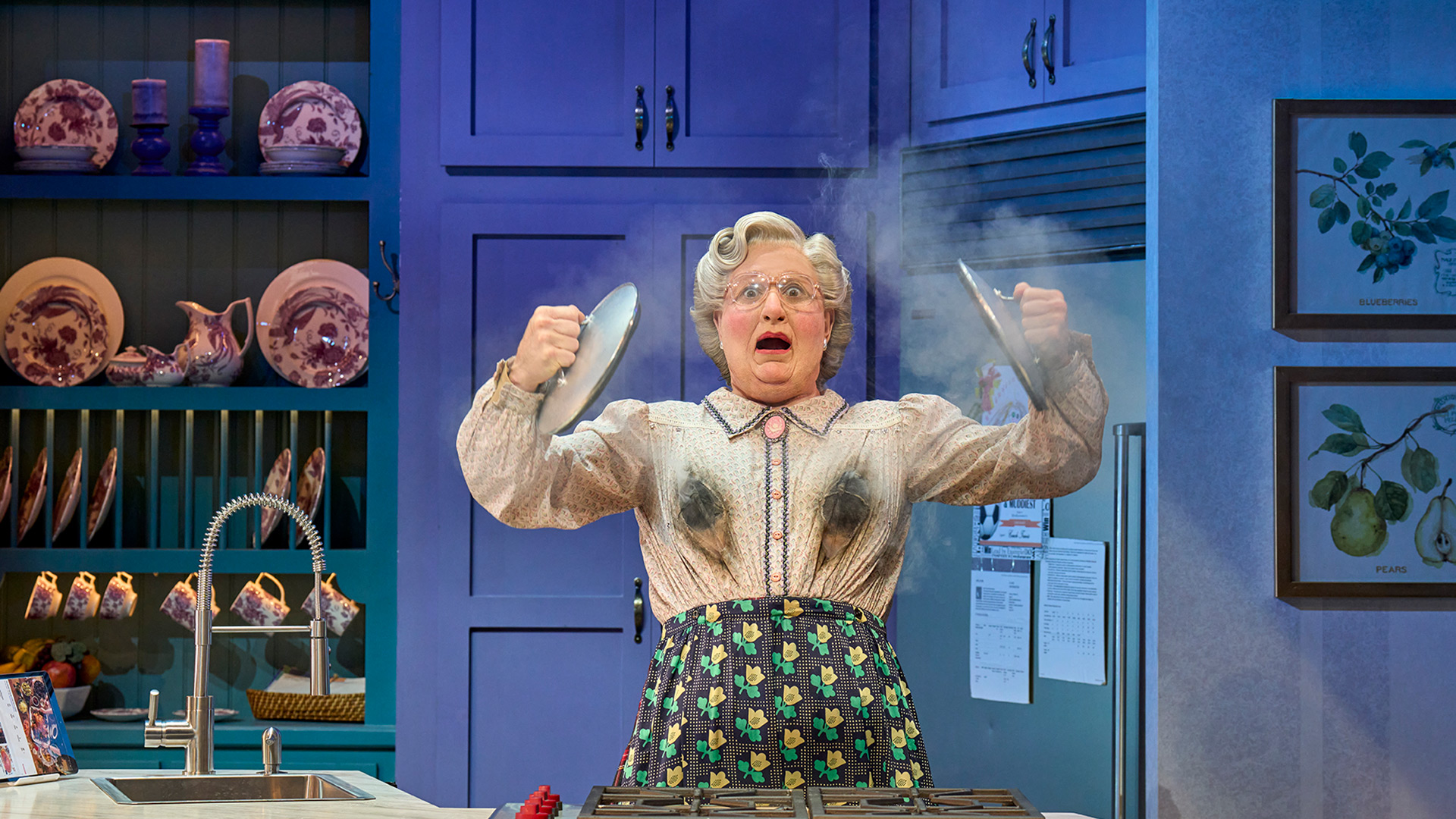 Laugh the night away with Mrs Doubtfire at the Shaftesbury Theatre. Image courtesy of Storyhouse PR/ photo credit: Manuel Harlan. Scottish nanny mrs doubtfire stands on stage holding two pot lids with a smoking bosom.