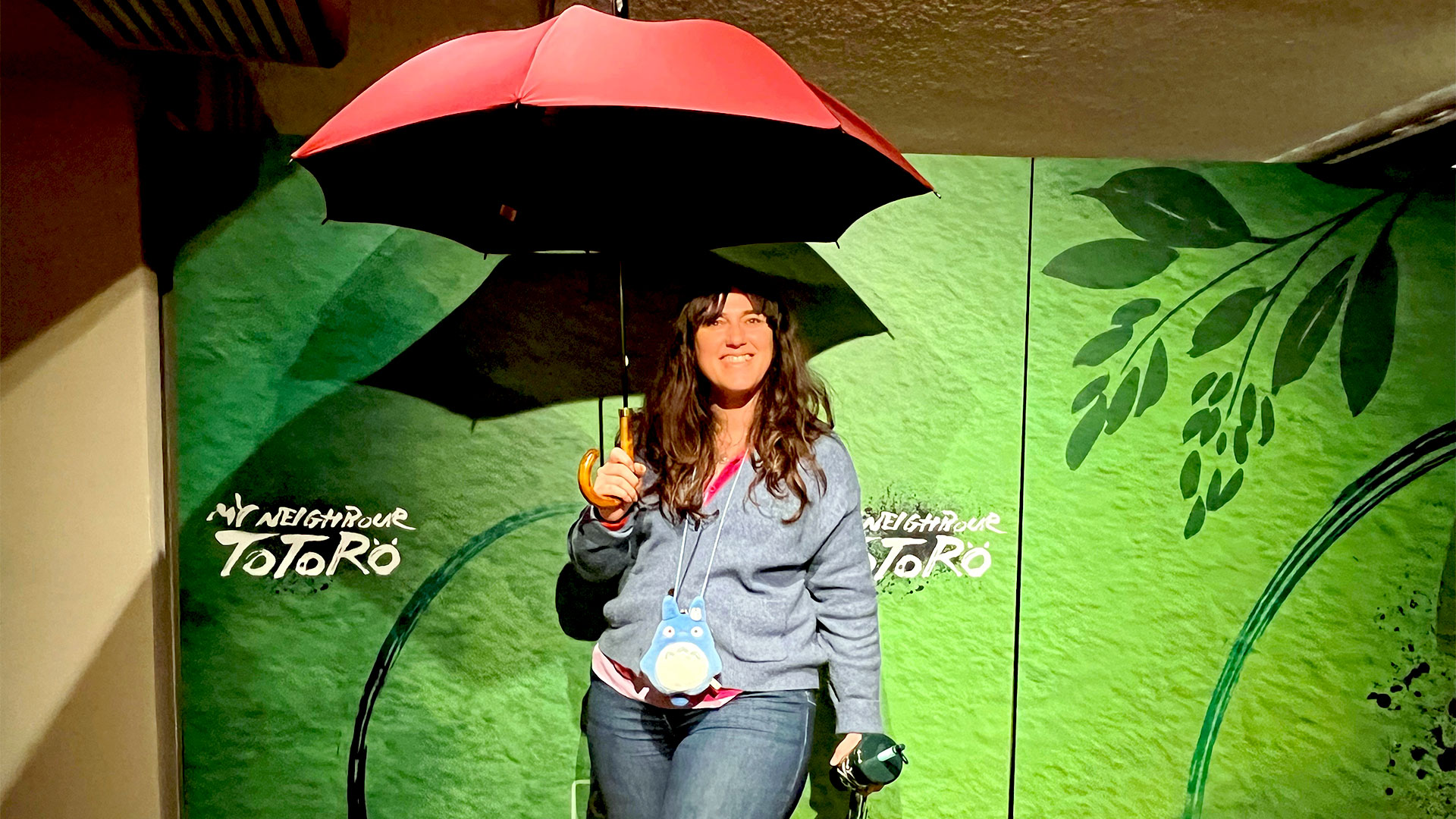 A woman stands under a red umbrella against a green backdrop, which is part of the stage adaptation of My Neighbour Totoro at the Gillian Lynne Theatre.