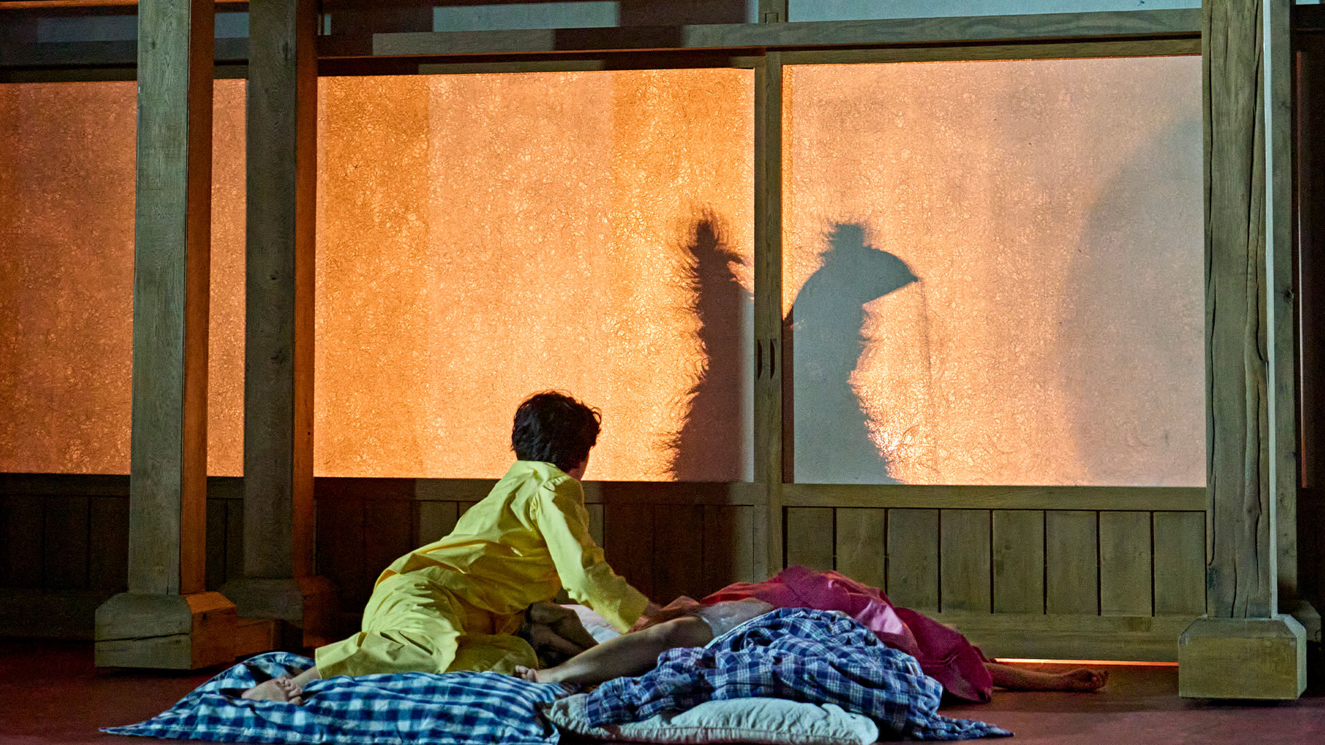 Satsuki (Ami Okumura Jones) wearing yellow pajamas and looking at a shadow of Totoro as Mei (Victoria Chen) sleeps next to her. 