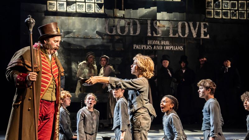 See one of the most popular musicals of all time return to the West End as Oliver! brings Dickens' story to life. Credit: Johan Persson. Image courtesy of Story House PR. A young boy playing Oliver holds a bowl over the Artful Dodger and asks for more gruel, while other children stand in the background.