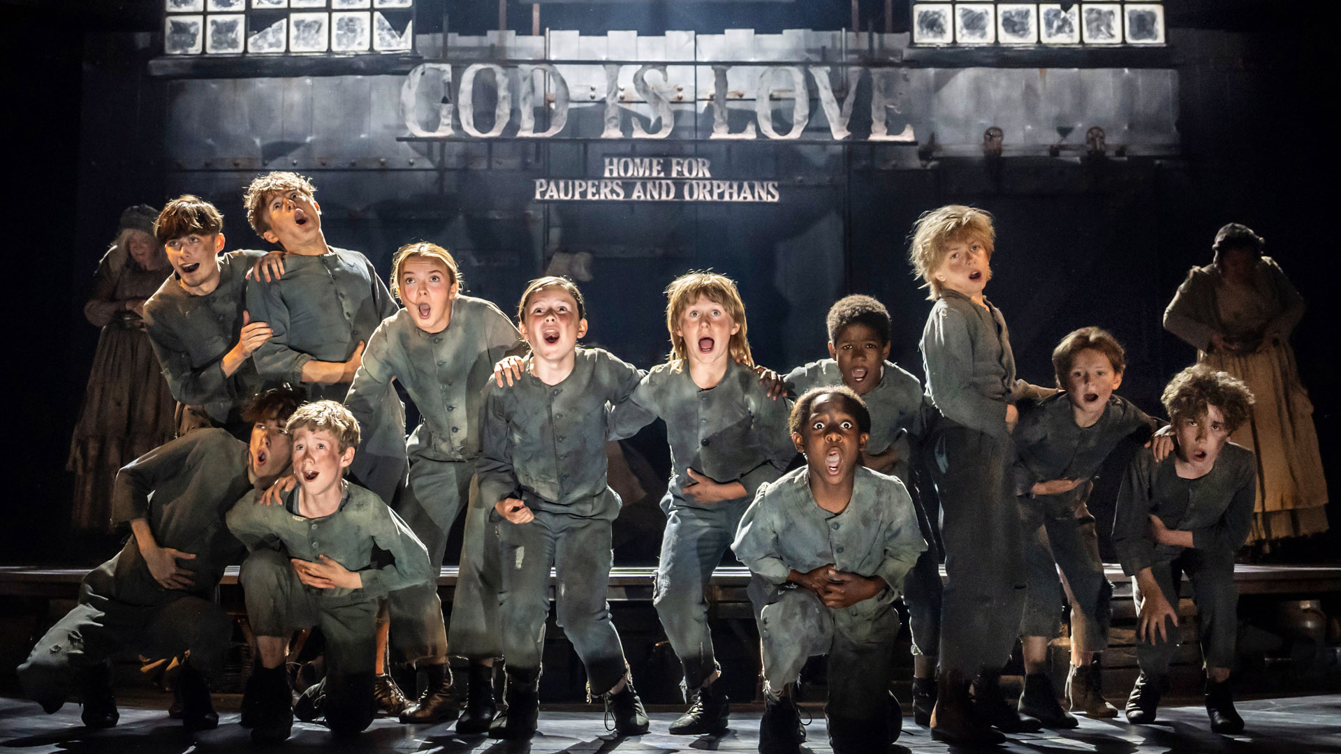See one of the most popular musicals of all time return to the West End as Oliver! brings Dickens' story to life. Image courtesy of Story House PR/ photo credit : Johan Persson A group of children dressed in dark grey uniforms, portraying the orphans in Oliver! The Musical are standing and singing on stage.