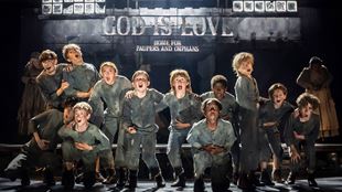 Eine Gruppe von Kindern in dunkelgrauen Uniformen, die die Waisenkinder in Oliver! The Musical darstellen, stehen auf der Bühne und singen.