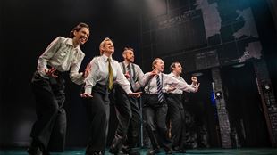 The five-person cast of Operation Mincemeat are standing and dancing on stage. They are all wearing black trousers, white shirts and ties.