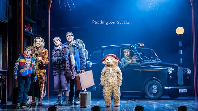Paddington Bear stands on stage next to the cast portraying the Brown family, with a black cab in the background at Paddington Station.