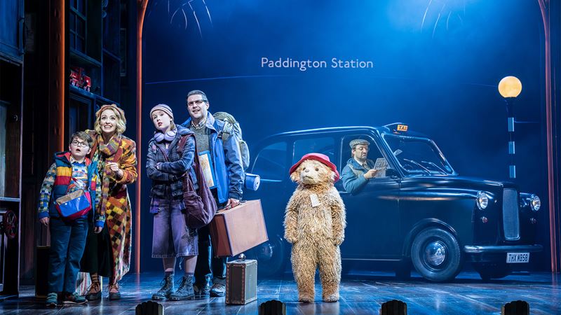 Paddington Bear stands on stage next to the cast portraying the Brown family, with a black cab in the background at Paddington Station.