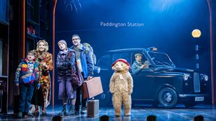Paddington Bear stands on stage next to the cast portraying the Brown family, with a black cab in the background at Paddington Station.