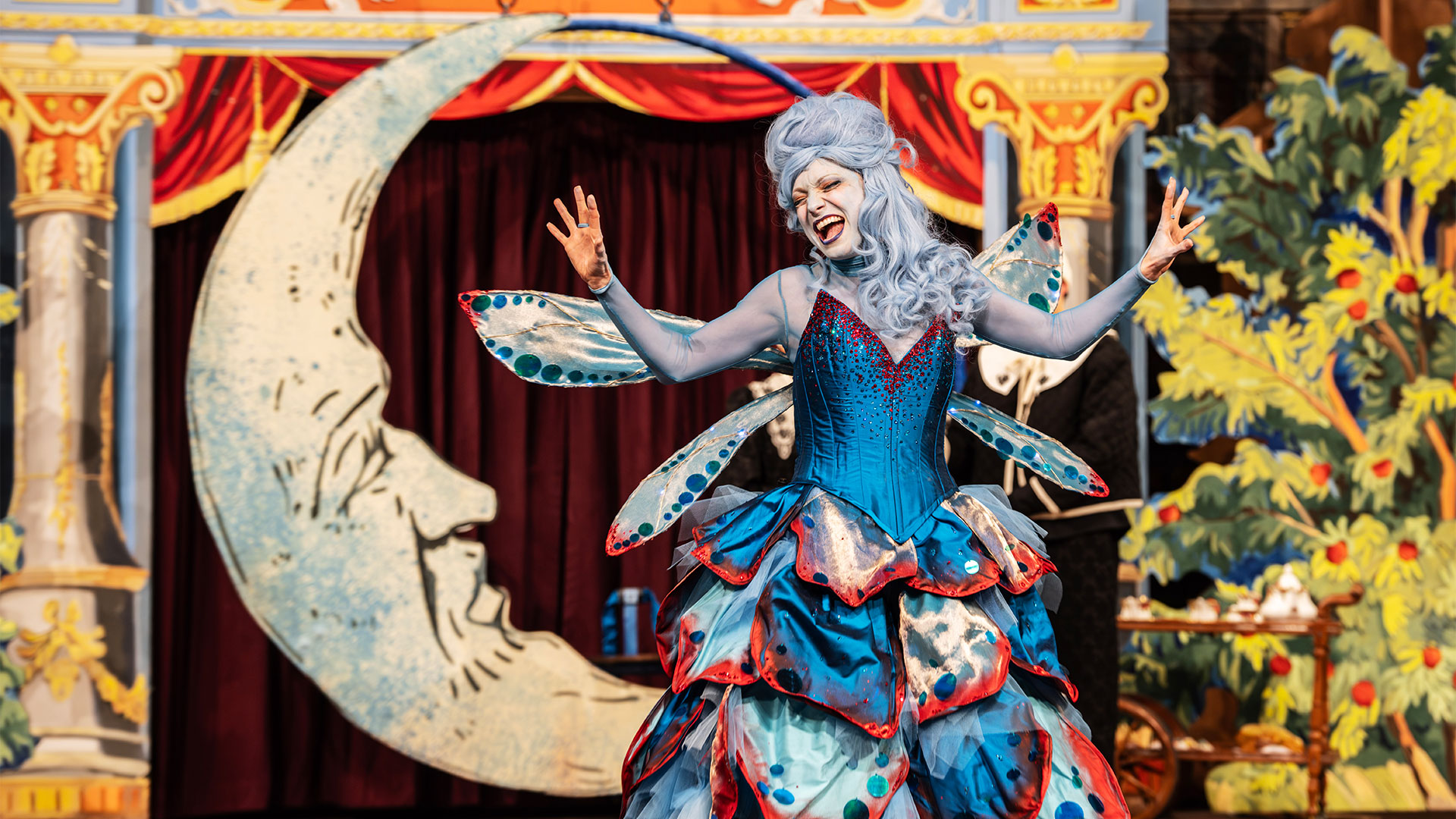 Tauchen Sie ein in die Welt von Pinocchio, in dieser neuen Bühnenadaption auf der Shakespeare Globe-Bühne zum Leben erweckt wird. Bildnachweis: Johan Persson. Als Blaue Fee im Musical Pinocchio gekleidet, trägt Lucy McCormick ein leuchtend blau-rotes Korsettkleid mit verschiedenen Flügeln.