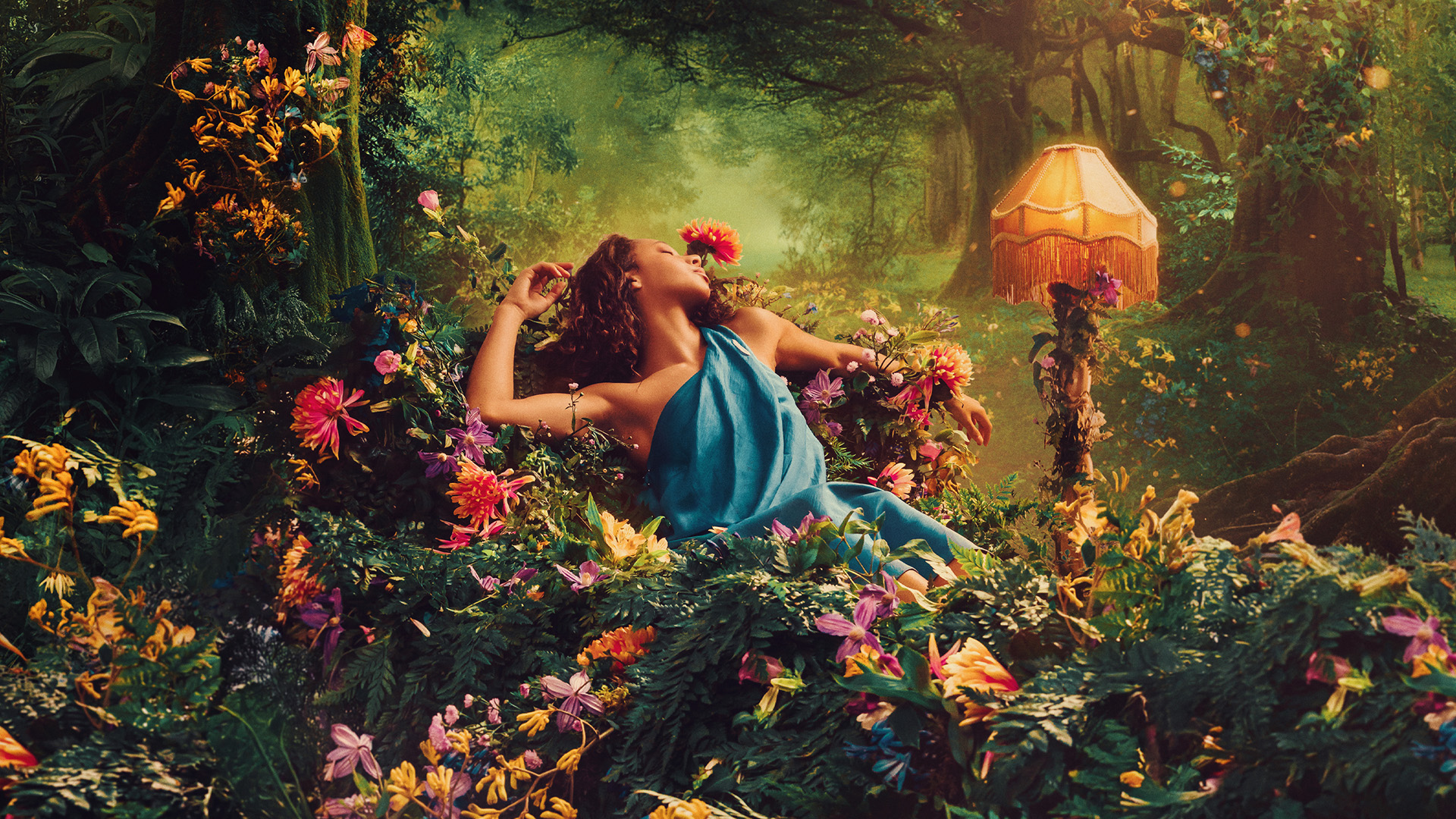 A young woman wearing a blue, draped dress lies down in a forest surrounded by colorful flowers and plants.