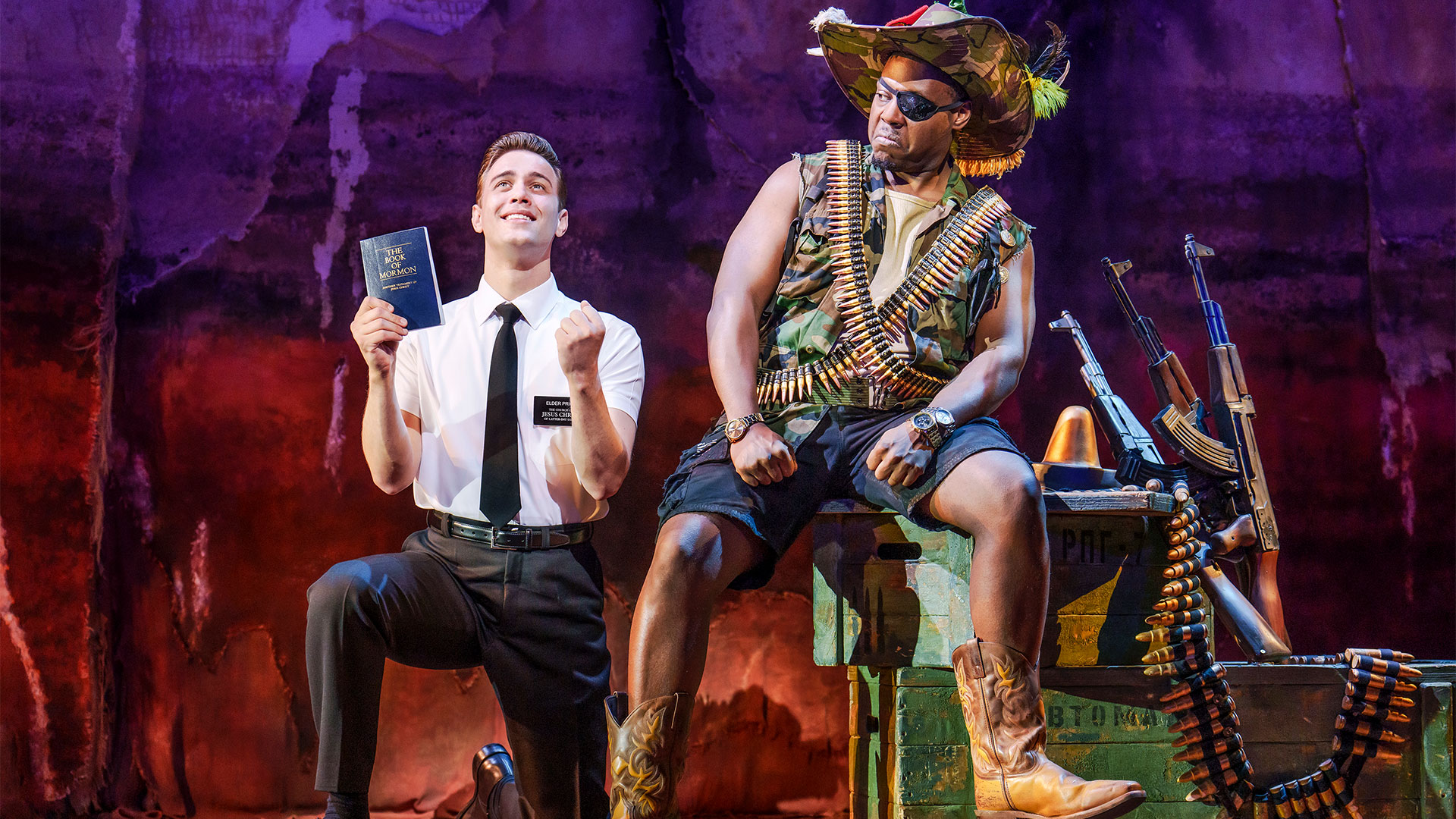 Join Mormon missionaries in a fun and humorous story when seeing The Book of Mormon in London. Image courtesy of London Theatre Direct. A missionary holding the Book of Mormon stands next to a man in camouflage, who is sitting on a box next to guns.