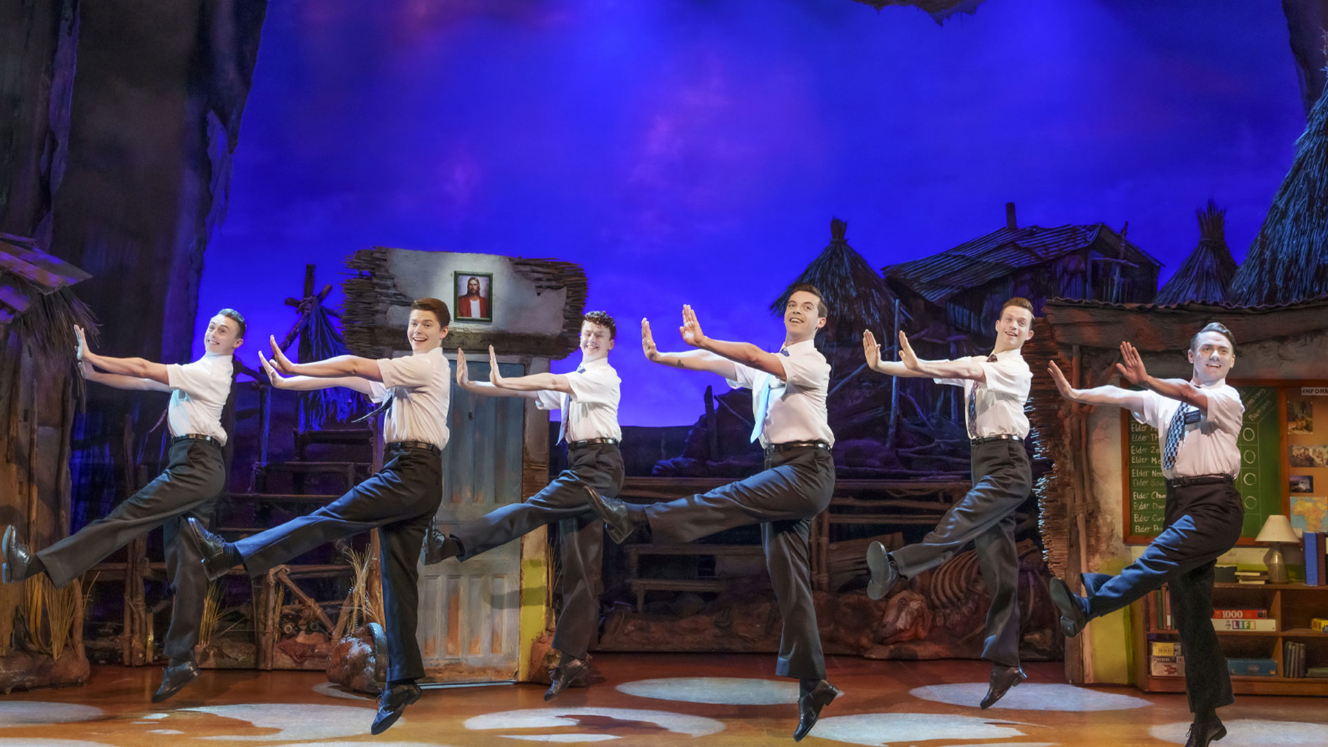 The cast of The Book of Mormon is dancing on stage in front of a dark blue background.