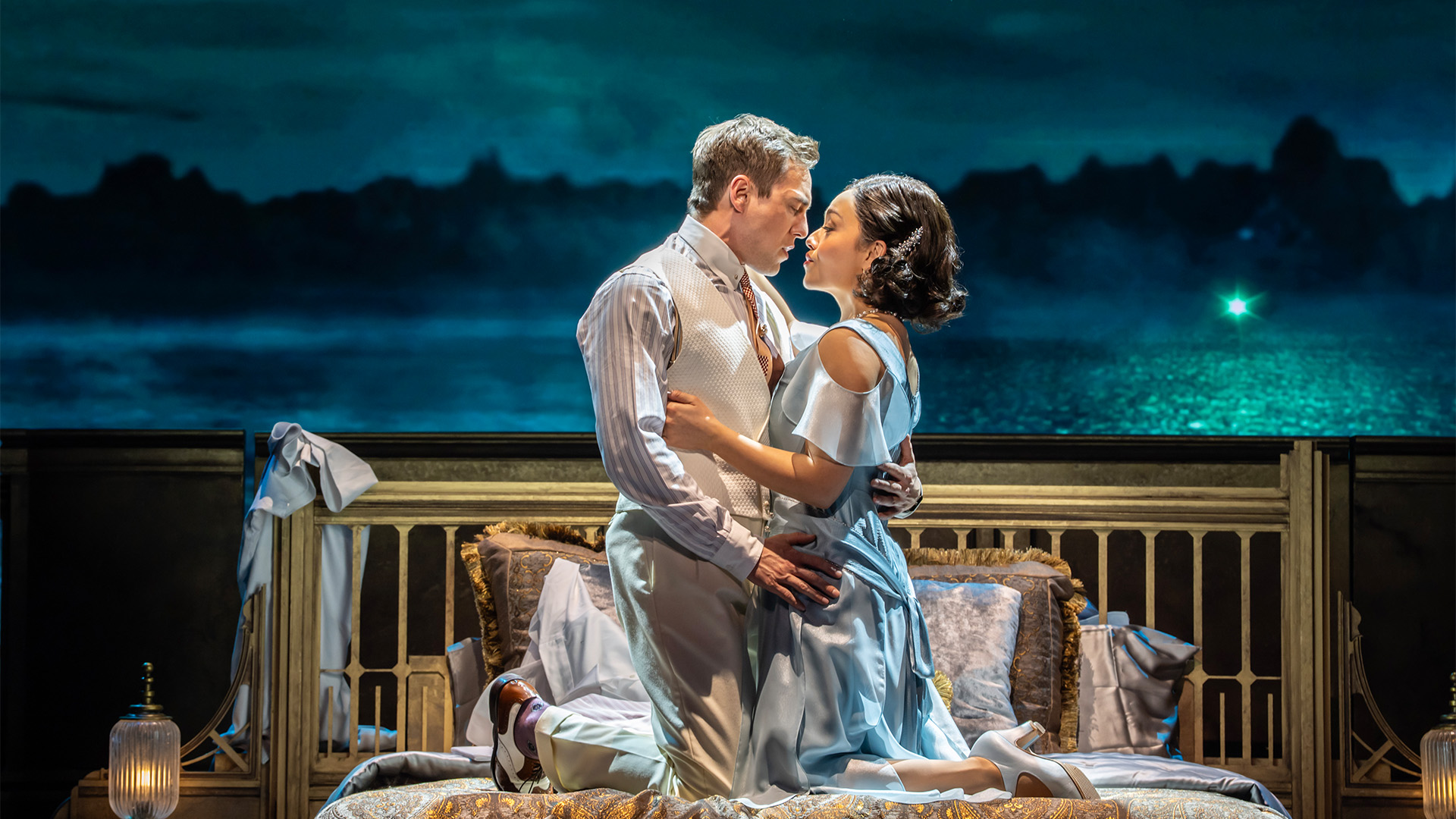 Two actors embrace on top of a bed against a coastal backdrop.
