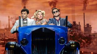 Three actors pictured in an old school car against a city backdrop in a West End show