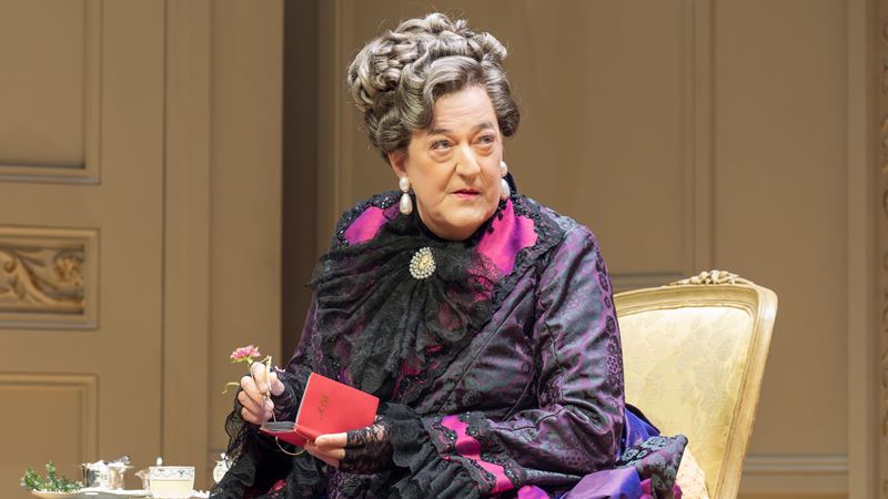 Actor stephen fry wears a black and pink dress and grey wig holding a red notebook in the importance of being earnest london theatre show.